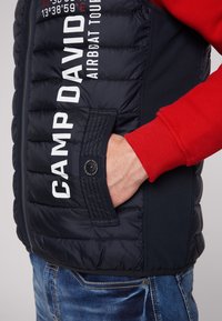 Gilet imbottito blu navy con sezioni trapuntate verticali, con testo bianco "CAMP DAVID" e una tasca laterale con bottone, abbinato a una camicia rossa a maniche lunghe.