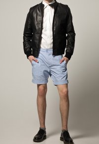 Svart läderbomberjacka med dragkedjor, vit skjorta, ljusblå shorts och svarta snörskor, stående mot en neutral bakgrund.