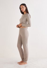 Femme debout pieds nus sur un tapis, portant un haut à manches longues beige et un pantalon assorti, regardant vers l'objectif avec une expression neutre.