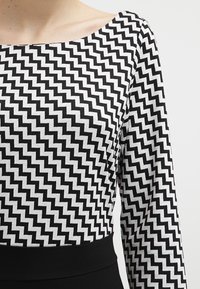 Haut noir et blanc avec un motif en zigzag, manches longues et encolure large, fabriqué à partir d'un tissu texturé. Ceinture noire.