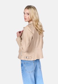 Beige leren motorjack met een aansluitend ontwerp, ritssluitingen en een ceintuurdetail op de taille, gecombineerd met lichtblauwe denim jeans.