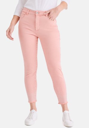 Frau trägt hellrosa enge Jeans mit weißen, funkelnden Details an den ausgefransten Saumenden am Knöchel, kombiniert mit weißen Turnschuhen und einem weißen Oberteil.