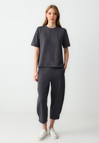 Donkergrijze katoenen blend-outfit met een kortmouwtop met decoratieve studs en losse broek met een omgeslagen zoom. Witte sneakers.