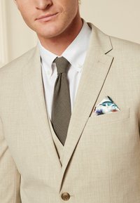 Chaqueta de traje beige con una textura sutil, combinada con una camisa blanca y una corbata gris con patrón. El pañuelo de bolsillo presenta un diseño floral.