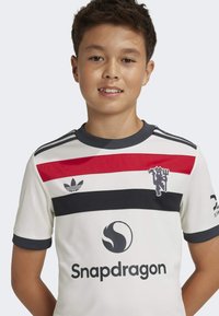 Camiseta branca de manga curta com riscas horizontais vermelhas e marinhas, apresentando um logo da Adidas e um gráfico do Snapdragon. Tecido texturizado.