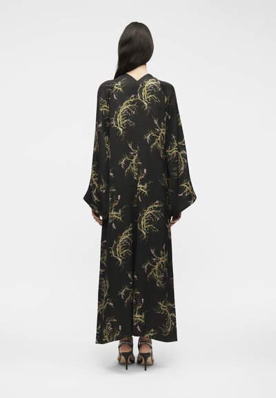 Γυναίκα με μακριά σκούρα μαλλιά που φοράει μαύρο μακρύ φόρεμα με φλοράλ μοτίβο, φαρδιά μανίκια και μαύρα ψηλοτάκουνα σανδάλια, εμφανιζόμενη από πίσω.