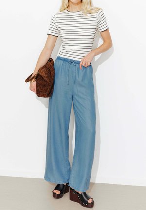 Femme portant un t-shirt rayé à manches courtes, un pantalon large bleu clair, des sandales compensées noires, tenant un sac marron à motif léopard.