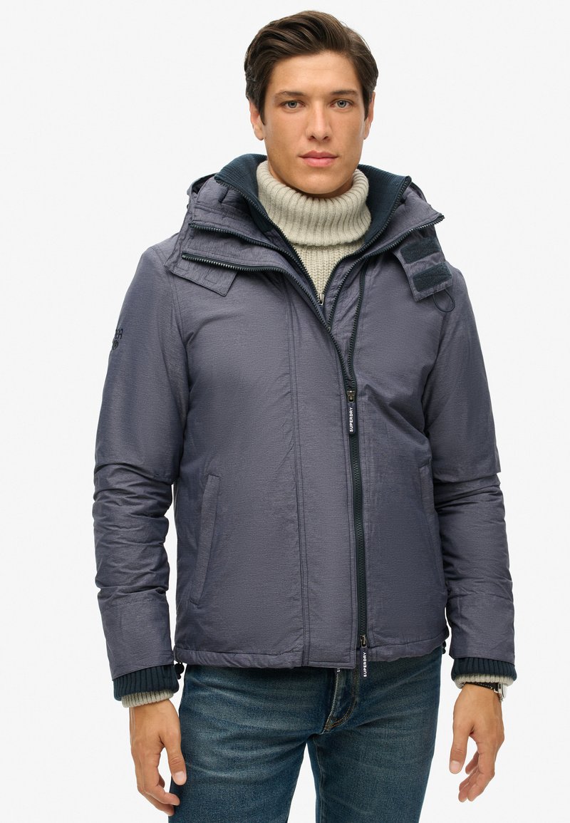 Granatowa puchówka z wysokim kołnierzem, zapinana na zamek, z bocznymi kieszeniami. Posiada fakturowany materiał oraz odpinany kaptur.