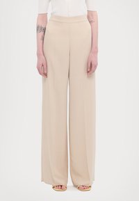 Pantaloni beige a gamba larga con una texture liscia, vita alta e senza hardware visibile. Design fluido che si estende fino a terra.
