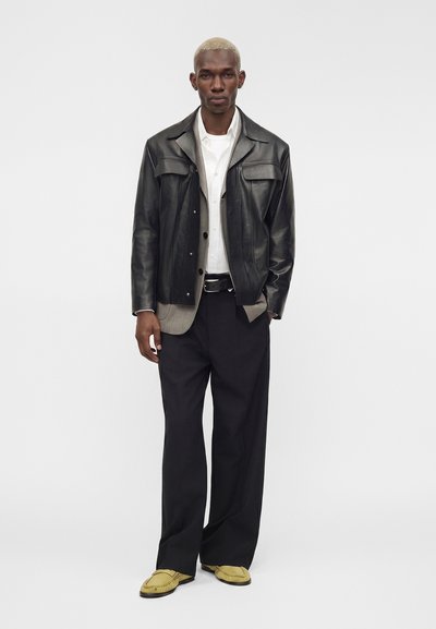 Homme aux cheveux blonds courts portant une veste en cuir noire par-dessus un blazer gris et une chemise blanche, un pantalon noir large et des chaussures marron clair.