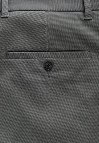 Poche arrière d'un pantalon en tissu gris avec une fermeture à un seul bouton noir et des passants de ceinture visibles en haut.