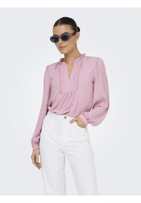 Blouse rose à manches longues avec un col à volants et des détails texturés, associée à un pantalon blanc taille haute. Accessoirisée avec des lunettes de soleil foncées.