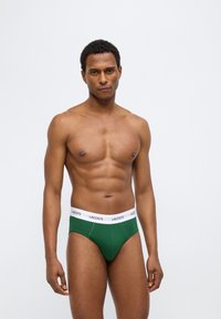 Πράσινα βαμβακερά εσώρουχα με λευκή ελαστική ζώνη μέσης που φέρει το λογότυπο "LACOSTE", παρουσιάζονται σε ανδρικό μοντέλο με ουδέτερο φόντο.