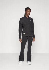 Svart zip-up träningsjacka med strukturerat mönster, kombinerad med matchande utsvängda byxor. Har en logotyp i ljus färg och sidofickor.