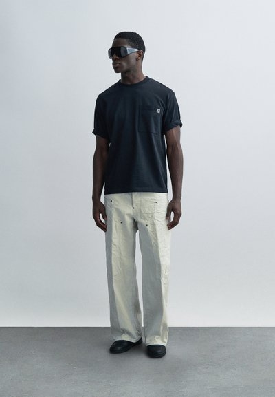 Camiseta de algodón negra con bolsillo en el pecho y mangas remangadas, combinada con pantalones cargo holgados de color gris claro y textura, y zapatos negros.