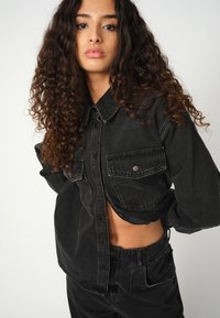 Schwarze Jeanshemd mit zwei Fronttaschen, Knopfverschluss und kastiger Passform. Über passenden schwarzen hochtaillierten Hosen getragen.