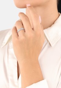 Silverring med sammankopplade band, med en rad små klara stenar längs toppen. Hand som försiktigt rör vid hakan, med ljus hy.