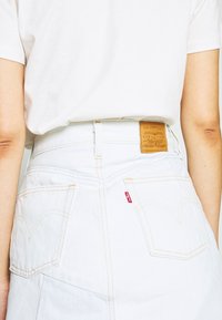 Bílá džínová sukně s vysokým pasem, žlutým šitím a zadními kapsami. Na zadní straně je značka z opálené kůže.