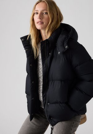Blonde Frau trägt eine schwarze Steppjacke über einem grauen Strickpullover, sitzt auf einem Holzstuhl vor einem einfarbigen hellen Hintergrund.