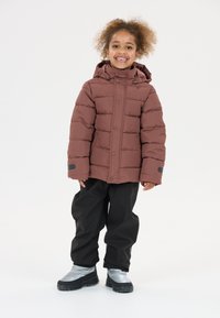 Doudoune marron avec capuche, présentant un design matelassé, associée à un pantalon noir imperméable et des bottes argentées imperméables.