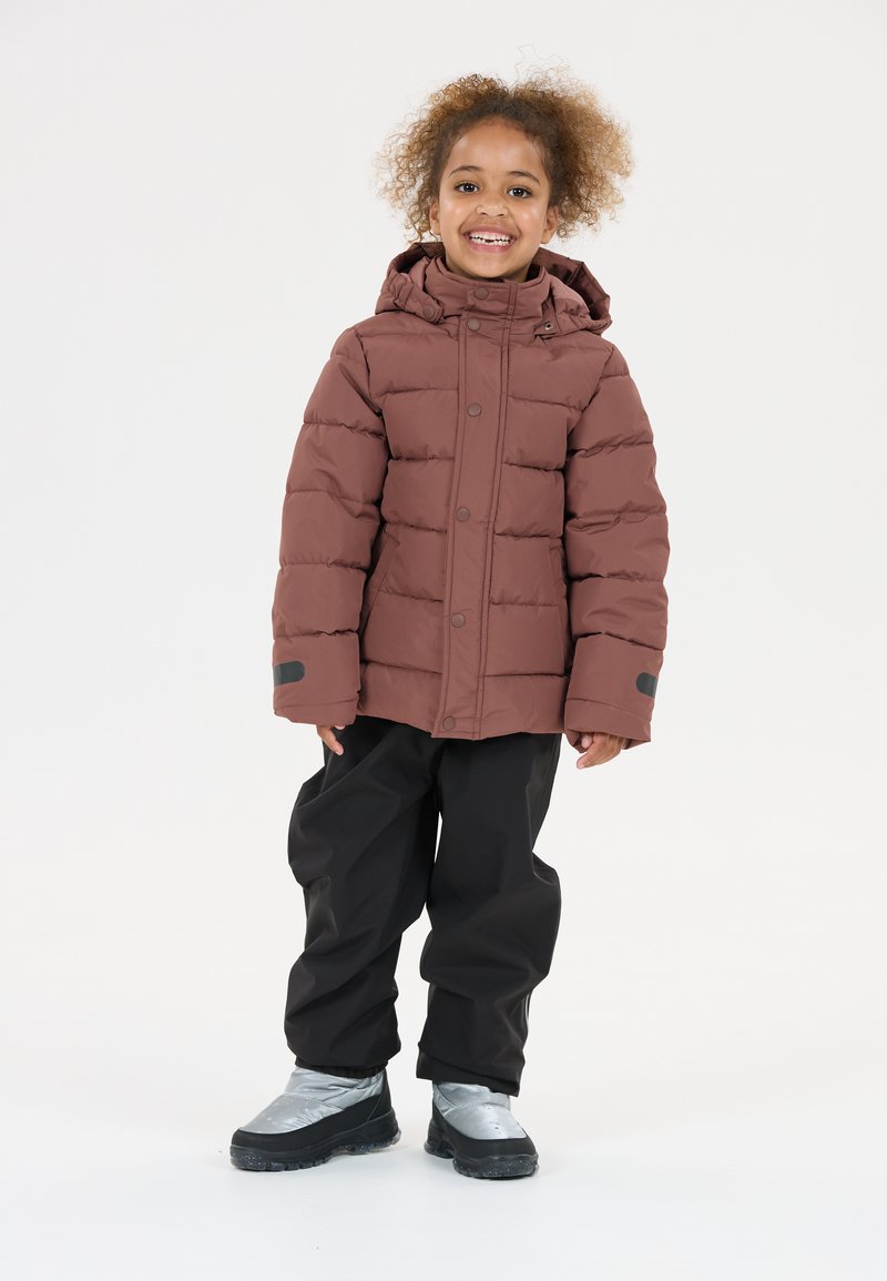 Doudoune marron avec capuche, présentant un design matelassé, associée à un pantalon noir imperméable et des bottes argentées imperméables.