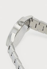 Reloj de pulsera de acero inoxidable plateado visto de lado, mostrando la corona y la pulsera unida, sobre un fondo blanco.