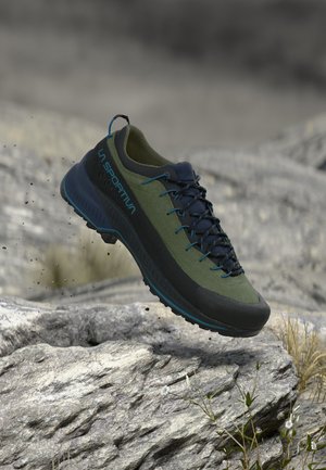 Olijfgroene en zwarte La Sportiva trailschoen met blauwe veters die zweeft boven rotsachtig terrein met kleine planten eromheen.