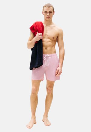 Svømmedragter i pink og hvidt ternet mønster med elastisk talje og snørre. Modellen holder et mørkeblåt og rødt håndklæde over sin skulder.