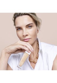 Foundation-flaske fra Lancôme holdt i en hånd, klar glass med et hvitt lokk, fylt med en lys beige væske. Bakgrunnen er nøytral.