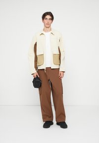 Beige and brown color-block jacket with two front pockets, paired with brown cargo pants and black sneakers. Small black bag worn on wrist.