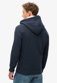 Granatowa bluza z kapturem, wykonana z miękkiego materiału, z kieszenią kangurką i ściągaczami przy mankietach. Prosty krój o luźnym fasonie, bez widocznych logo.