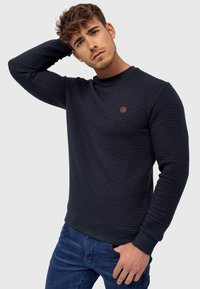 Navy textured sweatshirt with a round neckline and ribbed cuffs. Features a small brown leather logo patch on the chest. Worn with blue jeans.