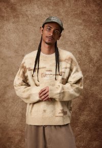 Ljusbeige ulltröja med ett strukturerat camouflagemönster och ribbade muddar. Bärs med en mjuk grön keps. Enkelt, avslappnat snitt.