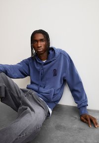 Sweat-shirt bleu à capuche avec une poche avant et un logo en ruban bleu foncé sur la poitrine, associé à un jean gris ample, le tout sur un fond neutre.