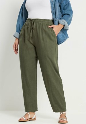 Kobieta ubrana w oliwkowozielone spodnie z troczkiem, biały top na ramiączkach, dżinsową kurtkę i białe sandały na paski na jednolitym tle.