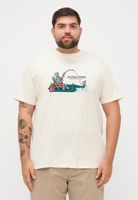 Beige Baumwoll-T-Shirt mit einem bunten Grafikmotiv eines Skeletts, das angelt, mit dem Markennamen "JACK & JONES" über dem Design.