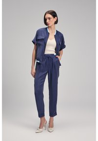 Mörkblå skjortjacka med två fickor och kontrasterande vit kant, i kombination med matchande smalbyxor. Modellen bär en krämfärgad topp och beige klackar.