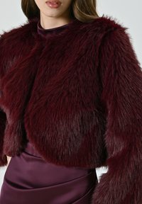 Giacca cropped in pelliccia artificiale bordeaux con superficie texturizzata e soffice e maniche larghe, abbinata a un vestito in satin coordinato.