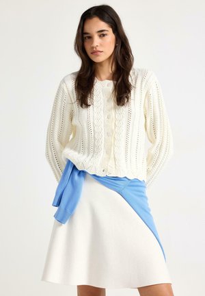 Giovane donna che indossa un cardigan a trecce bianco, una gonna bianca e un maglione blu legato in vita, su uno sfondo semplice.