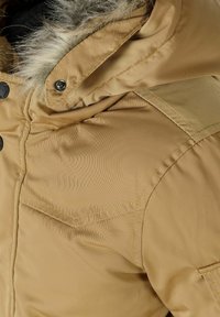 Giacca imbottita color tan con cappuccio rimovibile, presenta rifiniture in pelliccia sintetica, chiusura con bottoni a pressione e due tasche sul petto con cuciture in vista.
