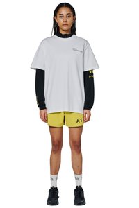 Młoda kobieta ubrana w oversize'ową białą koszulkę na czarnej koszulce z długim rękawem, żółte szorty, białe skarpetki i czarne trampki, stoi zwrócona przodem.