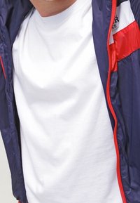 Námořnická modrá lehká bunda s červenými akcenty, zapínání na zip a bílým tričkem pod ní. Hladký materiál s sportovním designem.