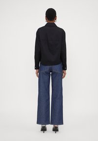 Camicia nera con colletto e maniche lunghe, abbinata a jeans in denim blu a gamba larga. Le scarpe hanno la punta affusolata e un tacco alto. Design minimalista.