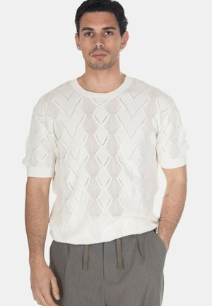 Uomo con capelli corti e scuri che indossa un maglione a maglia bianco a maniche corte con motivo e pantaloni grigi con le mani in tasca.