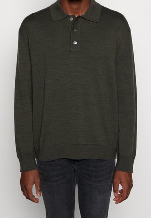 Mand iført en mørkeolivengrøn langærmet strikket polo med tre knapper, kombineret med sorte jeans, mod en ensfarvet hvid baggrund.