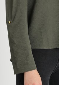 Blusa verde con texture e maniche lunghe, con dettagli di bottoni dorati sui polsini. Indossata sopra jeans scuri.