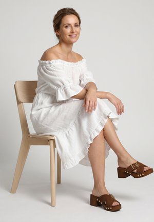Kvinde med lysebrunt hår i hvid off-shoulder kjole, der sidder på en træstol, smilende, iført brune platformsandaler med spænder.