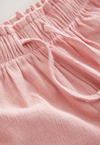 Světlounce růžová bavlněná látka s texturovaným povrchem, nařaseným elastickým pasem a stahovací šňůrkou. Sklady vytvářejí jemné záhyby.