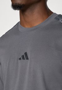T-shirt en coton gris avec un col rond, des manches courtes et un logo Adidas noir sur la poitrine gauche. Subtiles accents texturés sur les épaules.