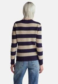 Osoba o krótkich platynowo blond włosach, ubrana w granatowo-beżowy sweter w paski oraz jasnoniebieskie jeansy, zwrócona tyłem na jednolitym tle.